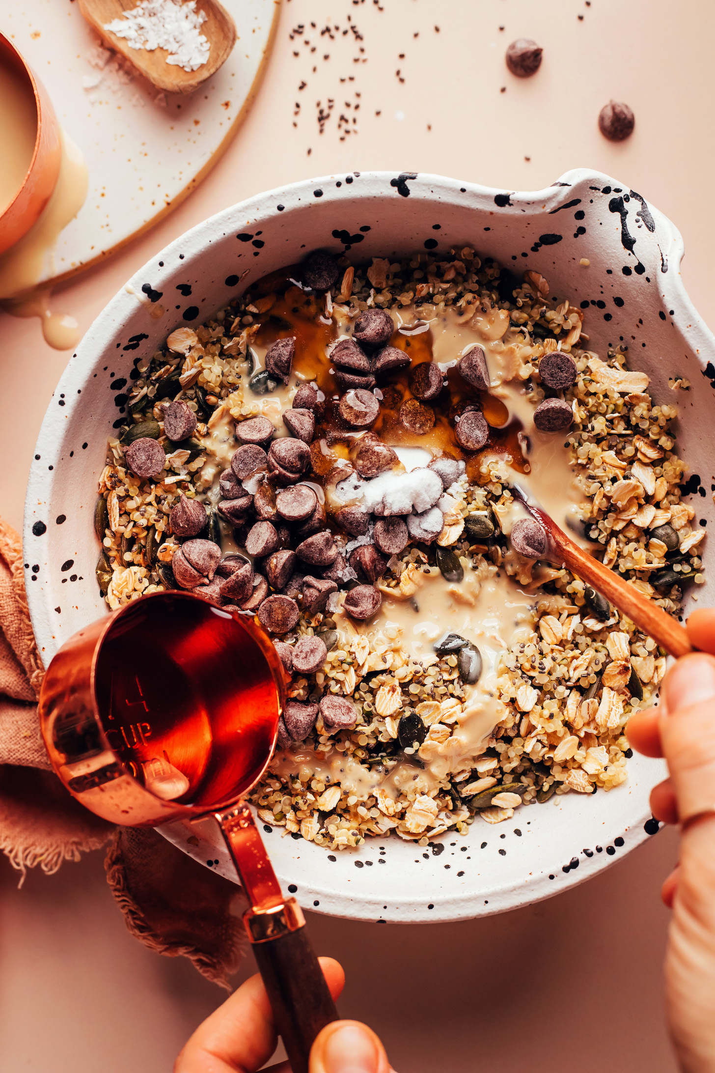 Pouring maple syrup into a bowl with quinoa, nuts, seeds, tahini, and chocolate chips